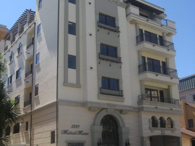 A multi-story beige building with balconies and arched entrance under clear blue sky.