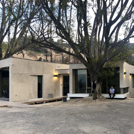 Small beige building near a large tree with people nearby in an outdoor setting.