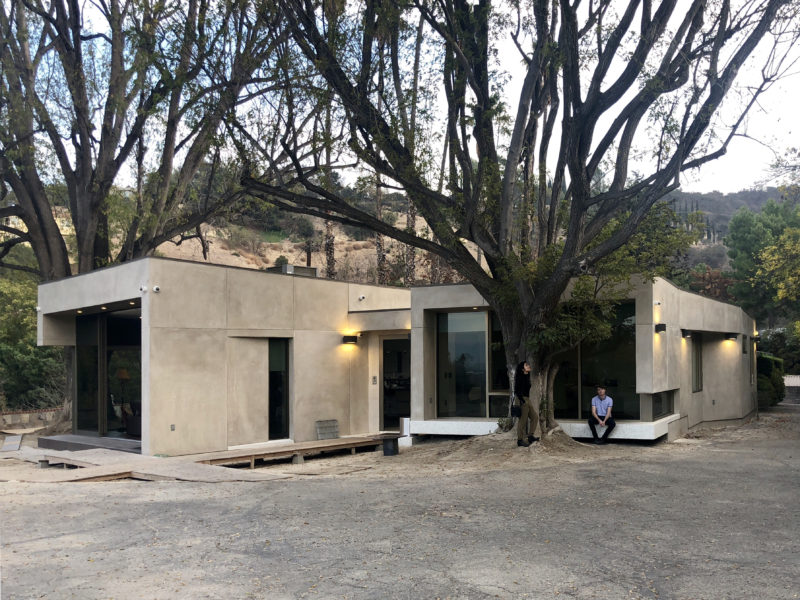 Small beige building near a large tree with people nearby in an outdoor setting.